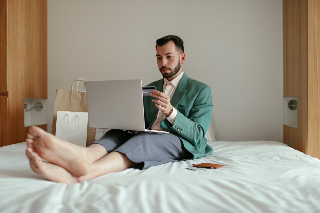 A man in a teal blazer shops online using his laptop and credit card while sitting on a bed.